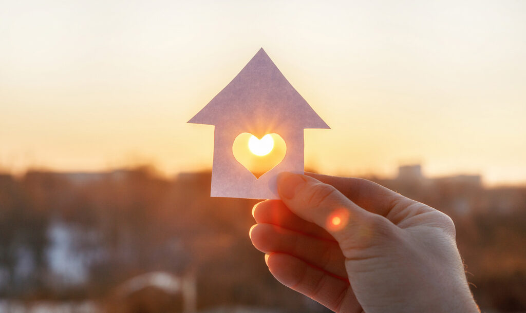 A hand holding up a paper house with a heart cut in the middle and the sun shining through.